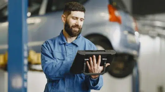 A maintenance technician in a blue uniform uses a rugged tablet to access DimoMaint MX on-premise software for real-time asset servicing in an automotive facility.