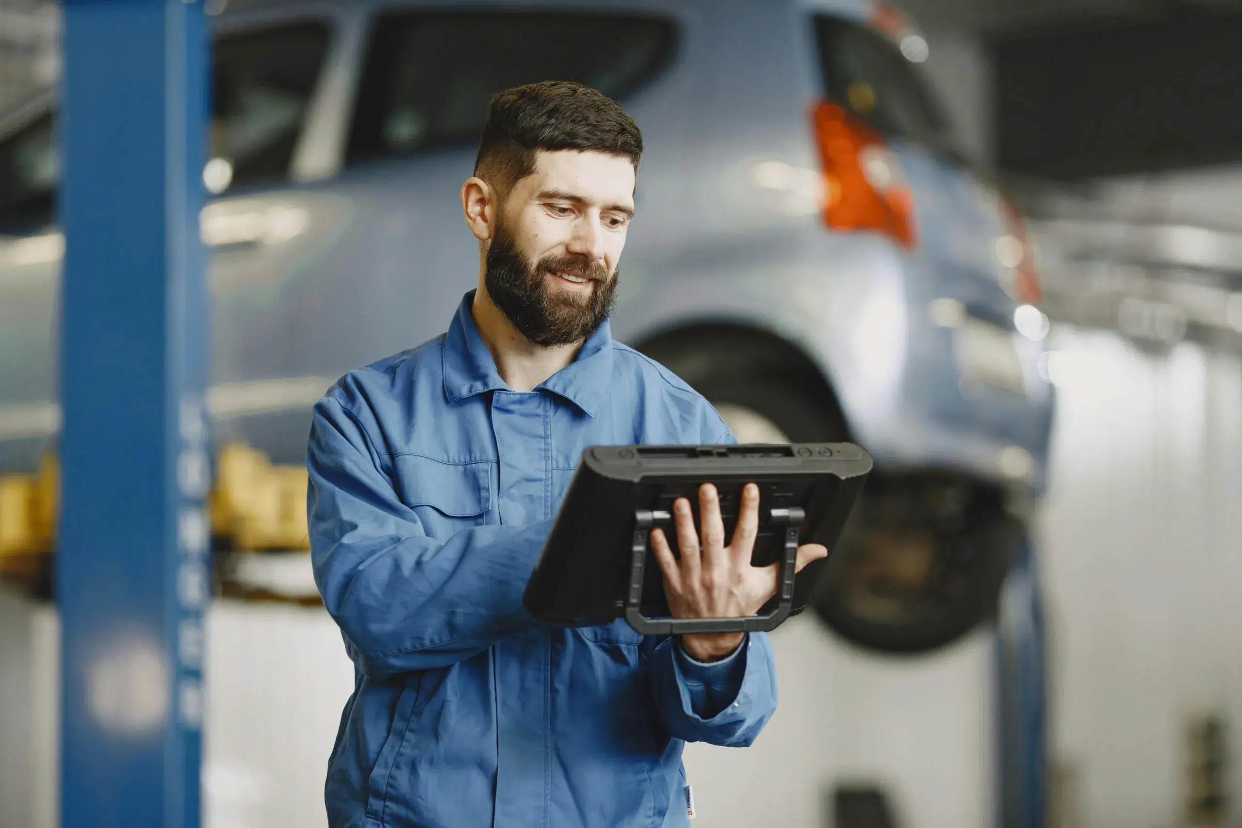 A maintenance technician in a blue uniform uses a rugged tablet to access DimoMaint MX on-premise software for real-time asset servicing in an automotive facility.