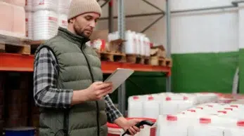 A logistics staff member using a tablet and barcode reader to perform a digital stock audit of palletized goods in a storage facility.