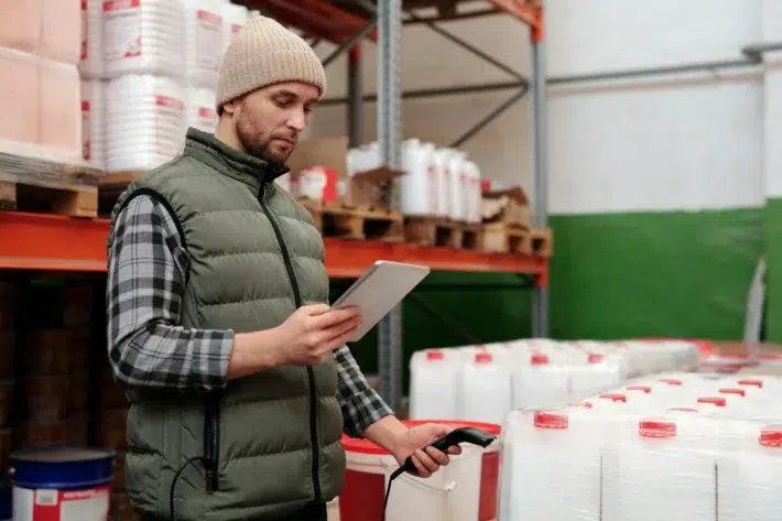 A logistics staff member using a tablet and barcode reader to perform a digital stock audit of palletized goods in a storage facility.