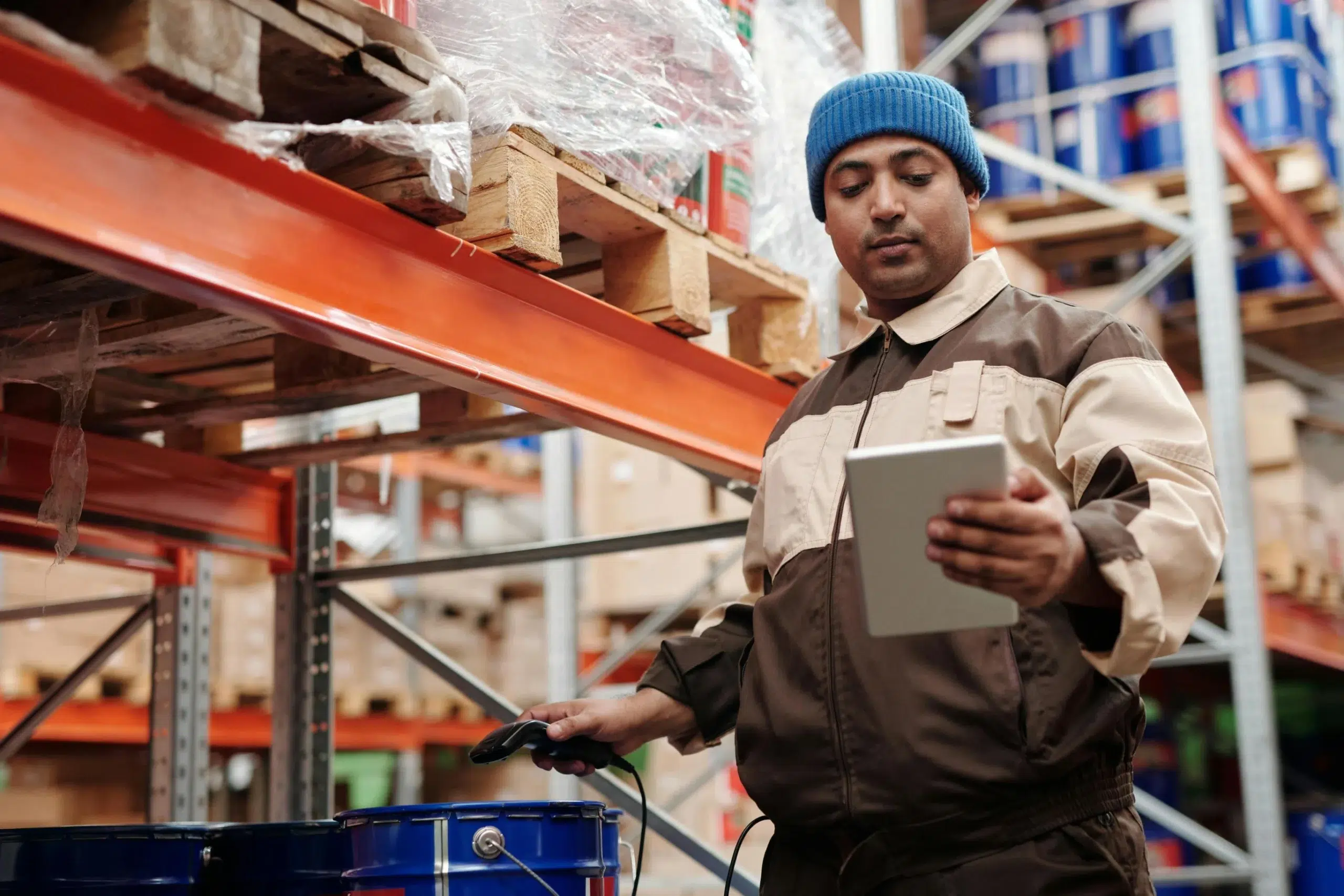 A warehouse technician in a blue beanie using a digital tablet and handheld barcode scanner to manage chemical drum inventory.