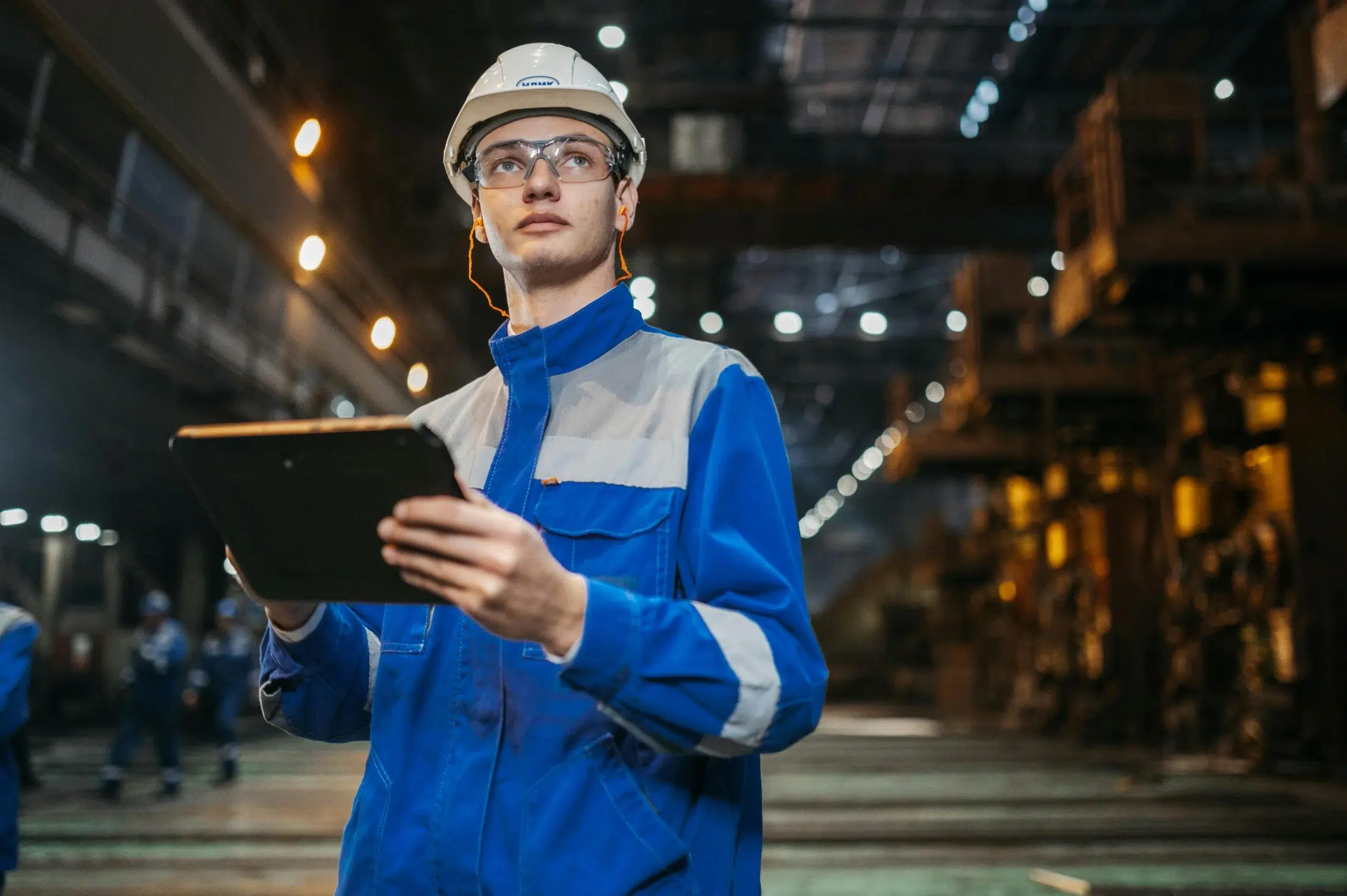 Industrial worker in blue uniform inspecting large-scale factory equipment using a mobile CMMS device for preventive maintenance checks.