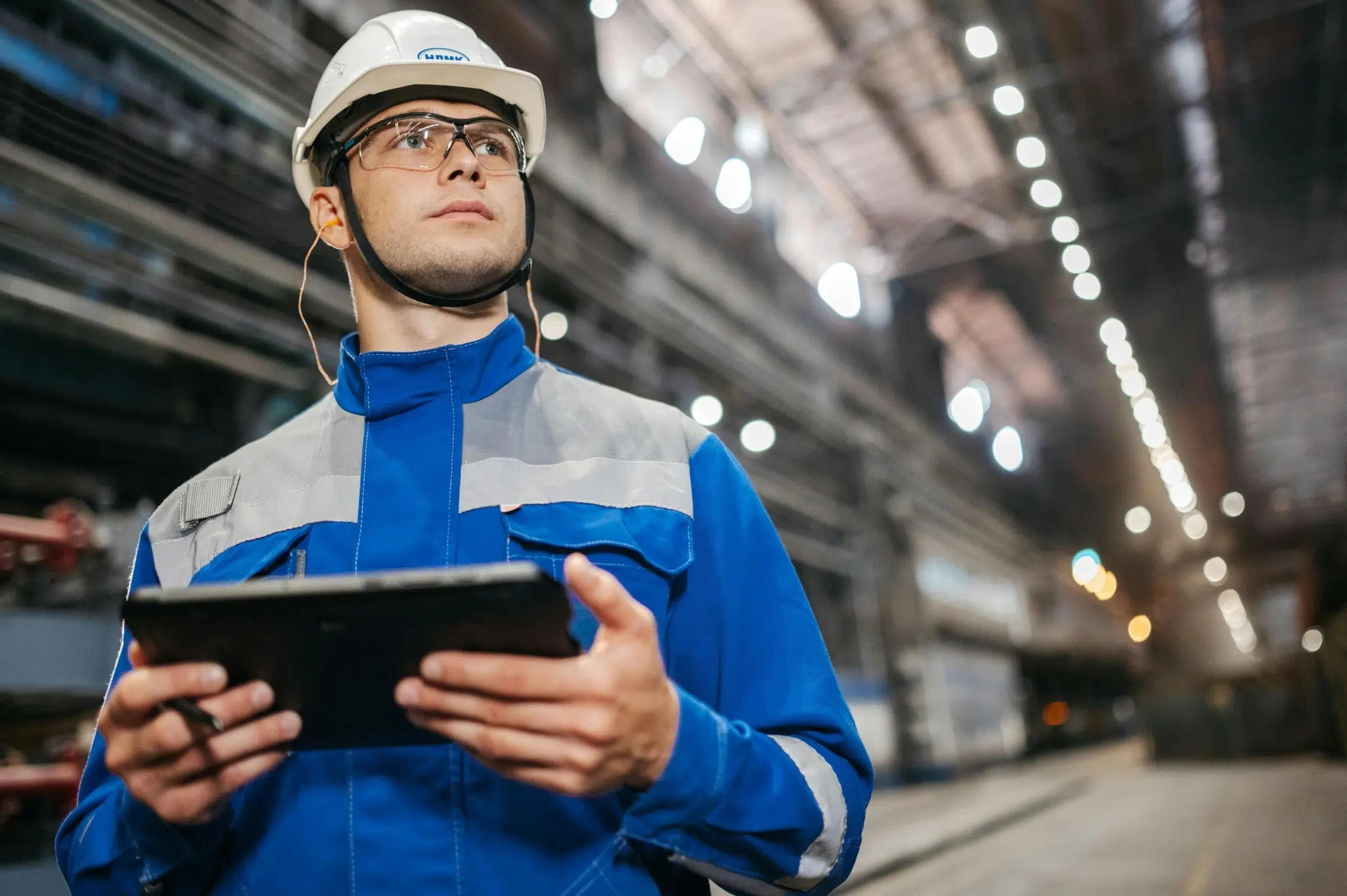 A maintenance technician in a white hard hat and safety glasses using a digital tablet to manage automated work orders in an industrial facility.A maintenance technician in a white hard hat and safety glasses using a digital tablet to manage automated work orders in an industrial facility.